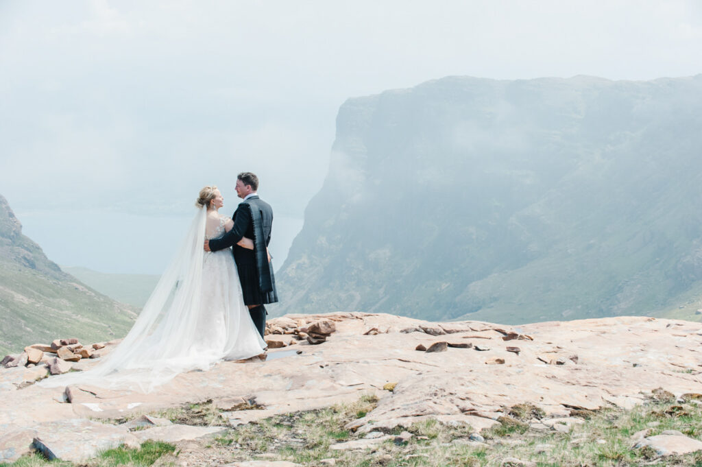 A married couple look at each other and embrace on a cliff top overlooking a large mountainous landscape