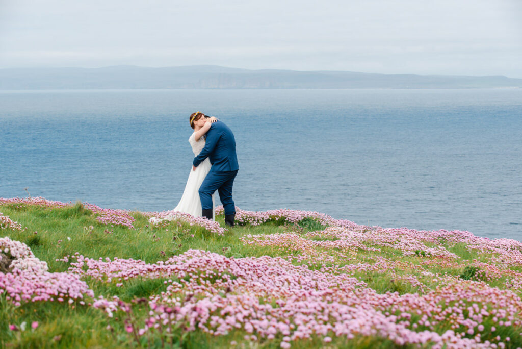 A newly married couple embrace on top of a cliff covered in pink sea thrift looking out to a large blue ocean and grey sky