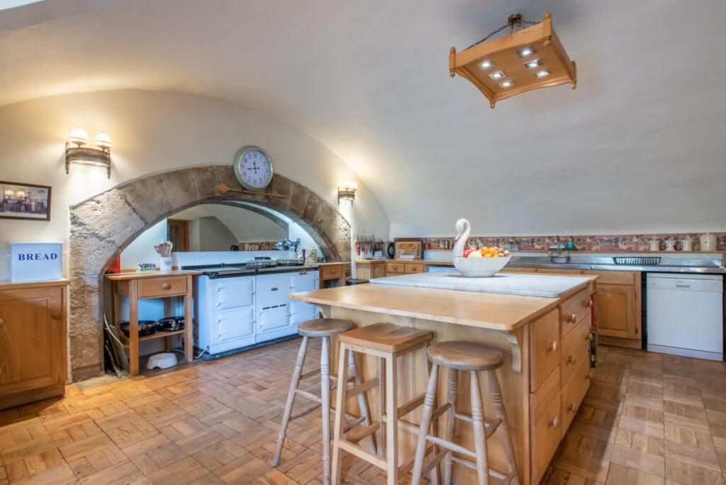 A white Aga stove sits beneath a stone arch in a a traditional kitchen with an arched ceiling and wooden island at its centre