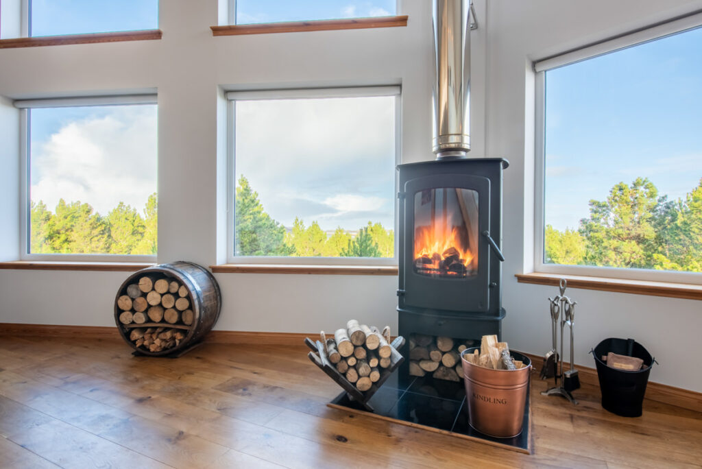 A modern upright log burner sits before three windows set in white walls in a room with wooden floors