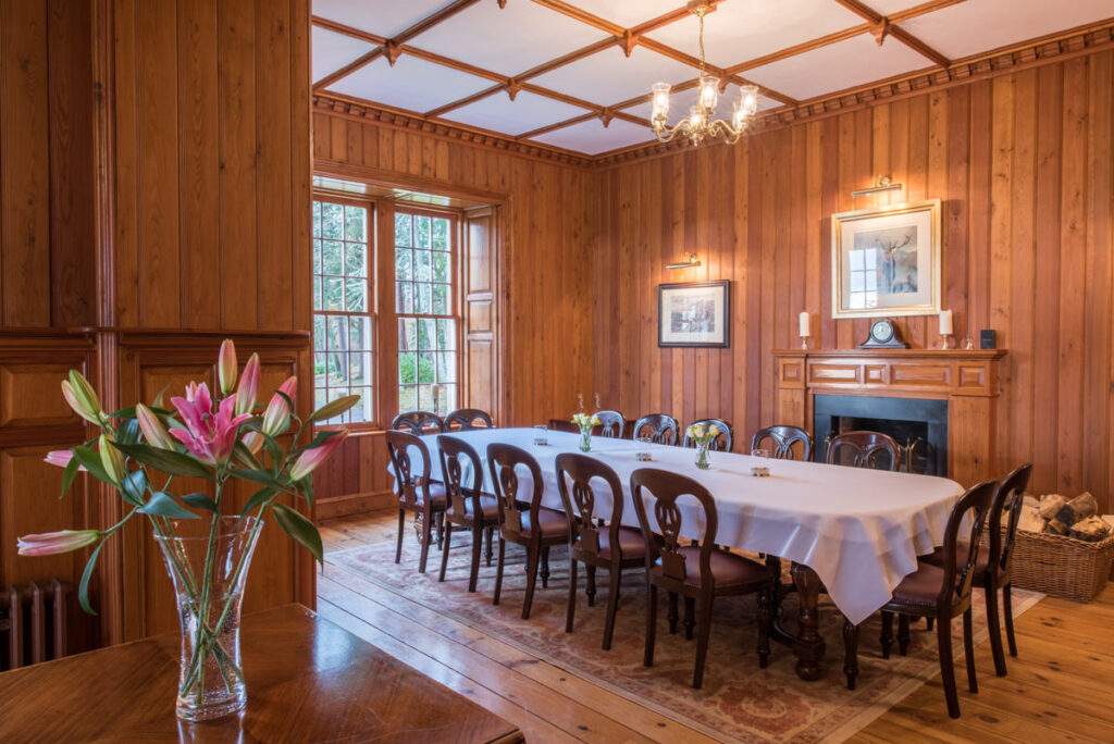 A large dark wooden dining table draped in a white table cloth sits below a Chandler in a wooden paneled room