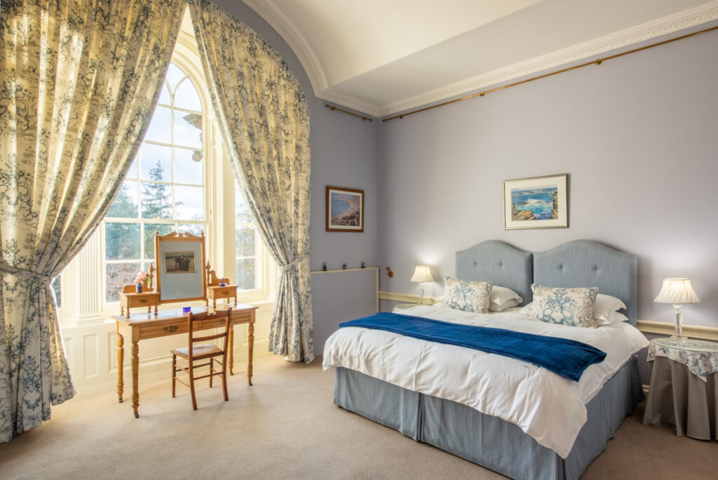 A twin bed sits in a large blue room with a large white window framed by blue floral curtains and dresser set at its centre