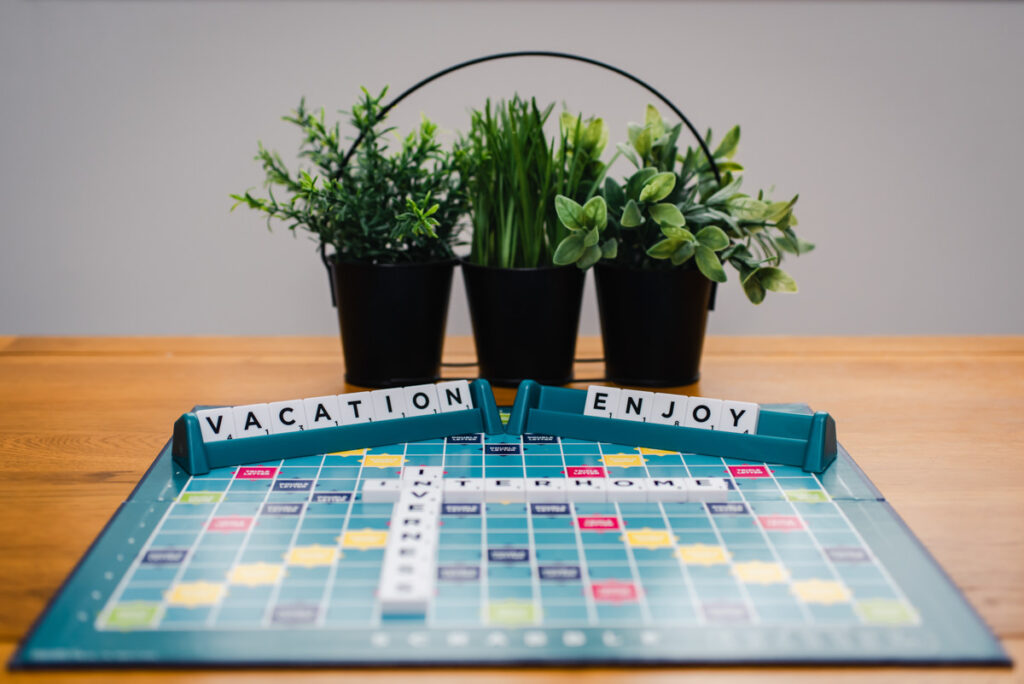 A close up a of a scrabble board on a pine table spelling out vacation enjoy with three black pots of herbs in the background