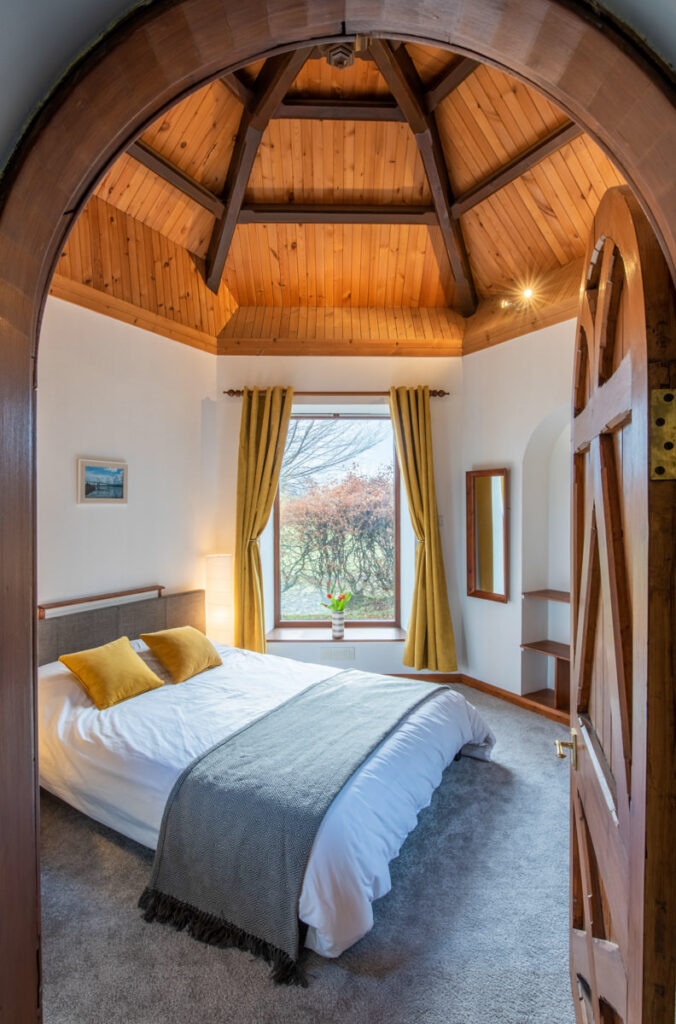 A view through an arched door into an octagonal room with a wooden pine ceiling, grey carpets, yellow curtains and double bed