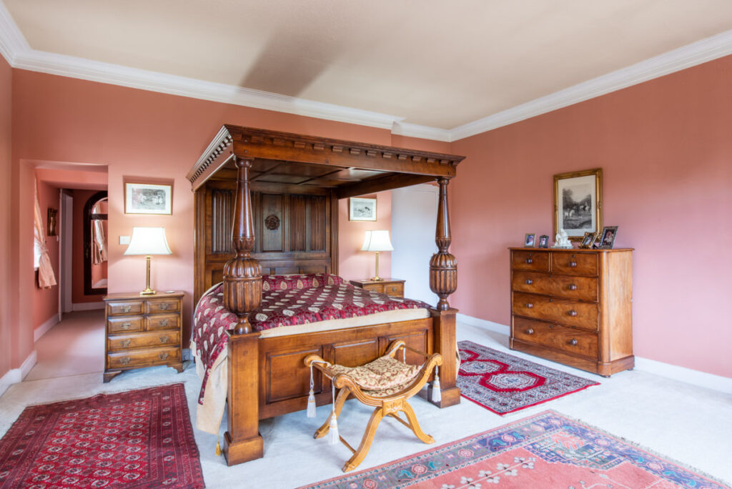 A large wooden four poster bed sits in the centre of a peach coloured room with rugs on the floor and wooden furniture