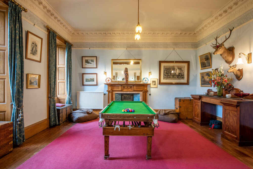 A snooker table sits on a big red rug in front of a fireplace in a traditional room with a stags head on a blue wall