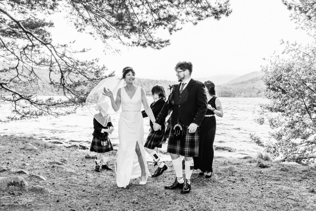 A monochrome image of a bride and groom holding hands next to a loch under some trees with the wedding party behind them