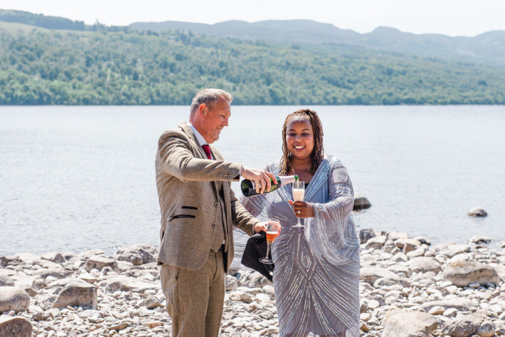 A mature couple share a glass of champagne while standing on a rocky beach by a Scottish loch on a sunny day