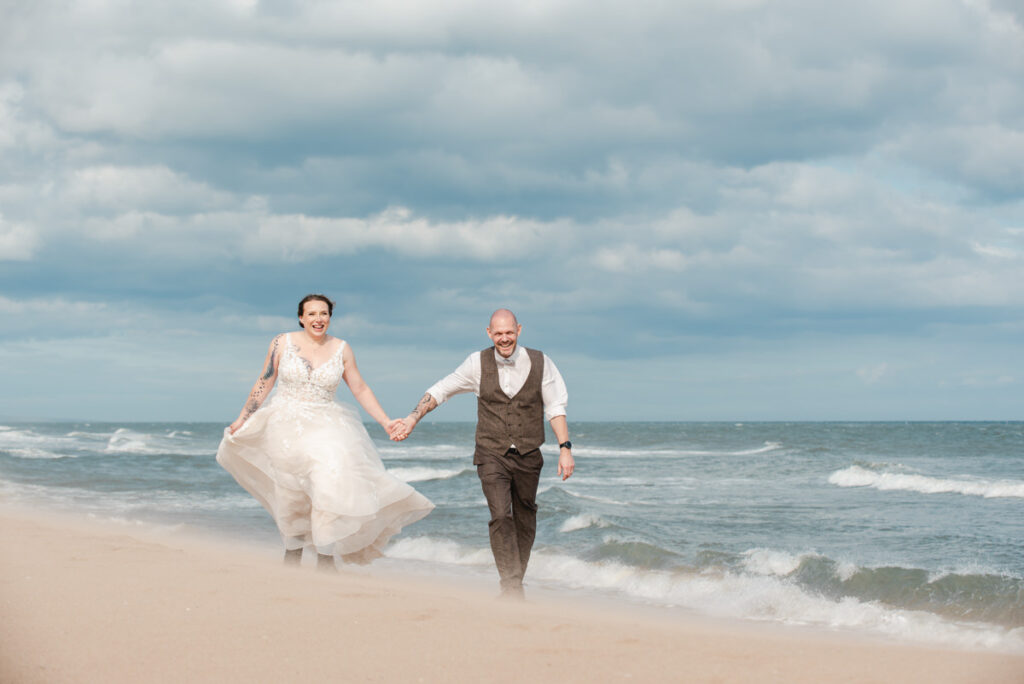A bride and groom hold hands while walking along a very windy and sandy beach with crashing waves to their left