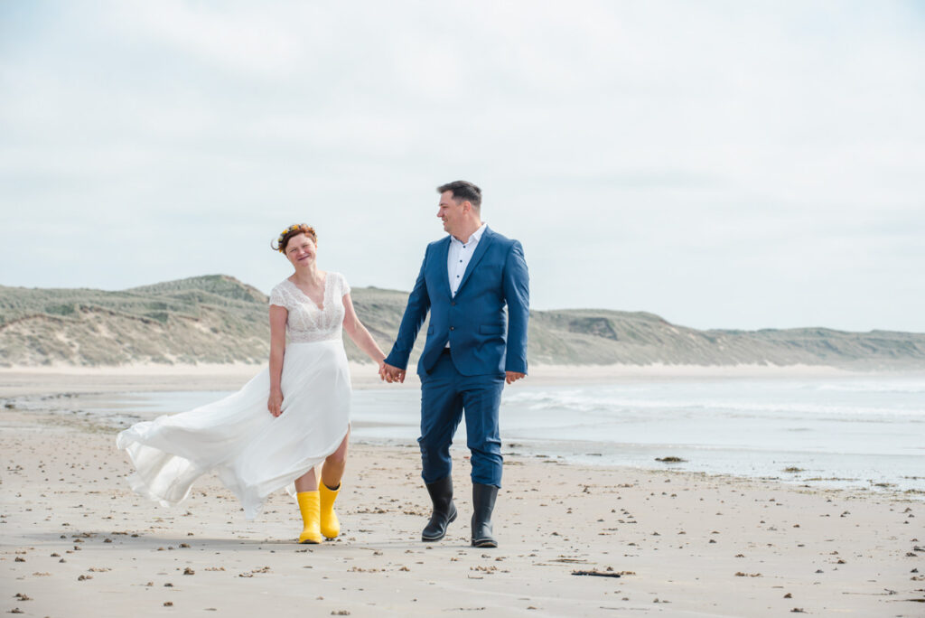 On a windy beach a bride in yellow wellies shares a joke while holding hands with her groom dressed in a blue suit