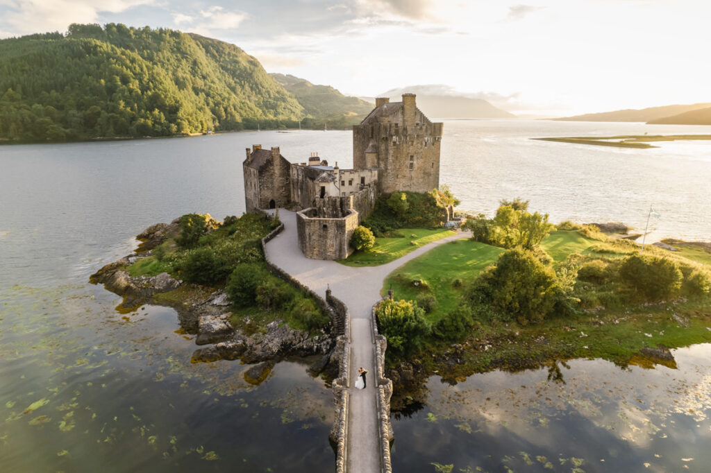 A drone shot of a couple standing on a bridge that leads to a large Scottish castle in front of a loch