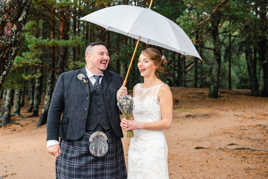 In a sandy, woodland setting, a kilted groom while laughing holds a white umbrella above his laughing brides head