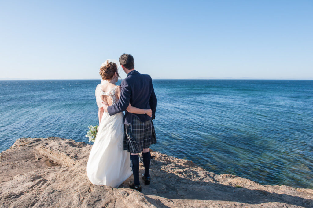 A newly married couple stand on a a stone clifftop while embracing and looking out to a large blue sea and cloudless blue sky