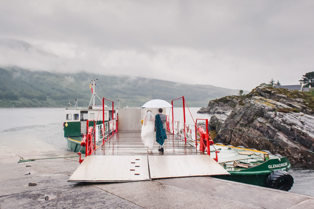 A bride and groom while holding a white umbrella walk onto a ferry during a very wet day on a Scottish island