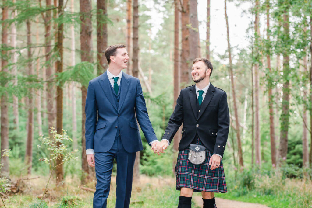 In a woodland setting a tall man in blue suit shares a joke with his husband who is dressed in a blue tartan kilt