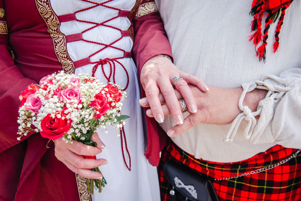 A close up of a bride and groom in red dress and red tartan holding hands to show off the wedding ring