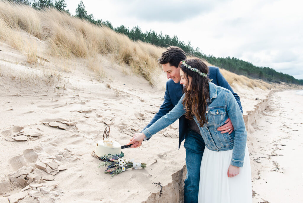 On a sandy beach a young newly married, casually dressed couple cut their wedding cake which sits precariously on a sand dune