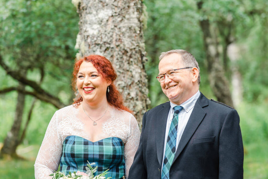 In a woodland setting a mature newly married couple stand together and smile with warmth at their out of shot wedding guests