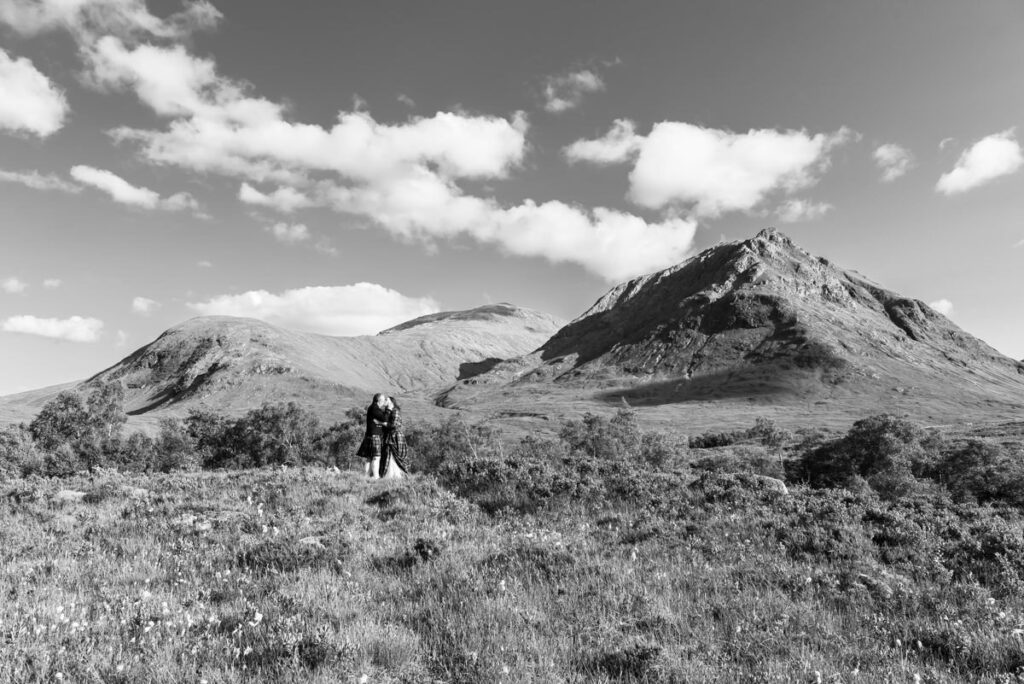 A monochrome image of a bride and groom embracing in the middle of a heather heath before a large mountain