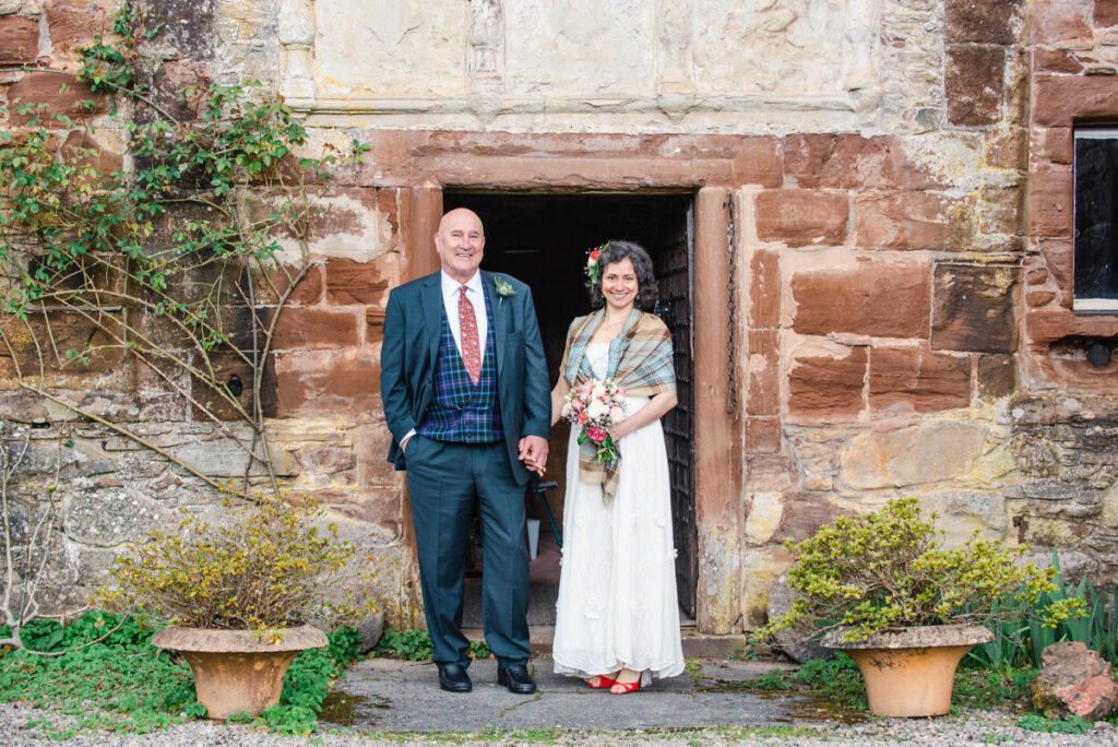 A newly wedded couple stand in an old sandstone doorway with a climbing rose on their right and terracotta pots at their feet