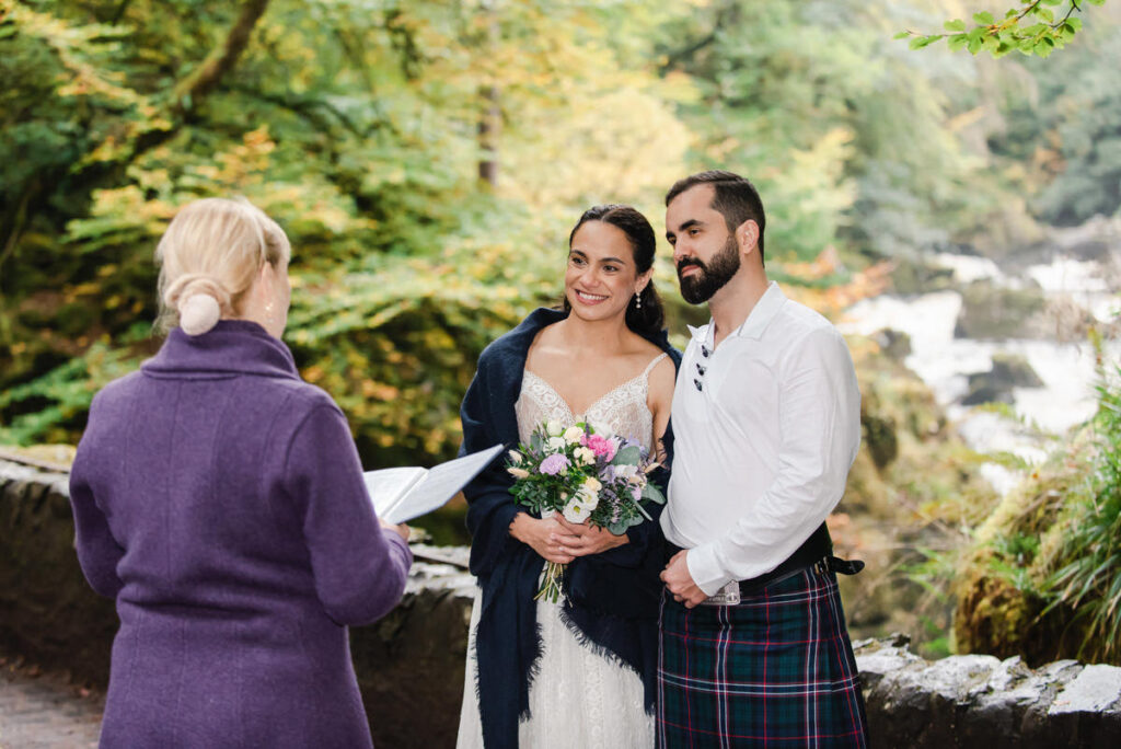 A celebrant in purple reads the wedding vows to a woman dressed in white with a blue shawl and bearded man in a kilt
