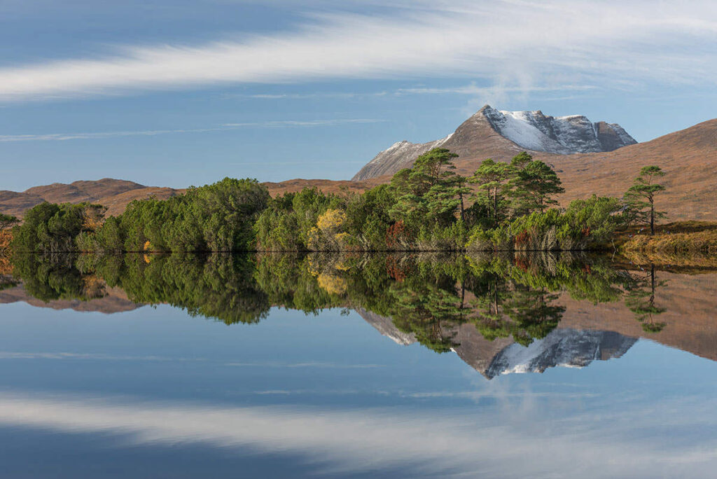 A reflection of a snow-capped mountain, green trees and blue sky with clouds in Coigach