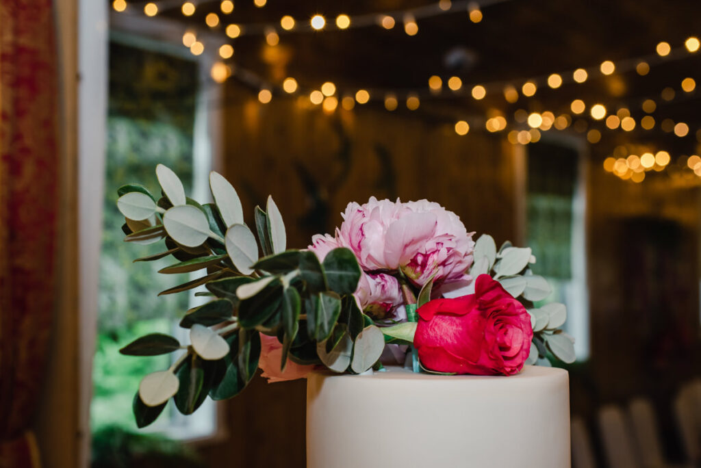 A wedding cake decorated with a red rose in a village hall with wooden walls and fairy lights